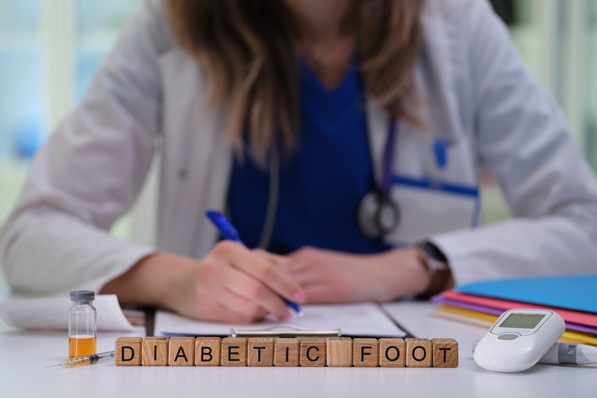 Ein Arzt schreibt an einem Schreibtisch, im Vordergrund sind Blöcke mit der Aufschrift „Diabetischer Fuß“ und medizinische Gegenstände zu sehen. - © H_Ko - stock.adobe.com