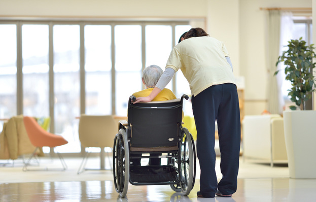 In einer hellen, modernen Pflegeeinrichtung stützt eine Pflegekraft sanft eine ältere Person im Rollstuhl. - © godfather - stock.adobe.com