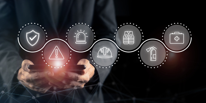 Eine Person hält ein Telefon mit Sicherheitssymbolen für Sicherheit, Notfall, Weste, Helm, Feuer und Erste Hilfe in der Hand. - © Parradee - stock.adobe.com