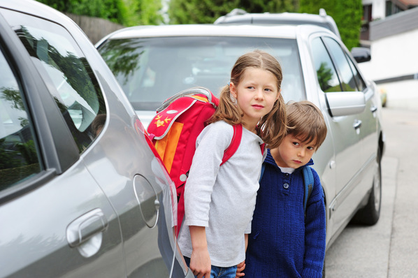 Zwei Kinder mit Rucksäcken stehen zwischen geparkten Autos auf einer Straße und schauen in die Kamera. - © photophonie - stock.adobe.com